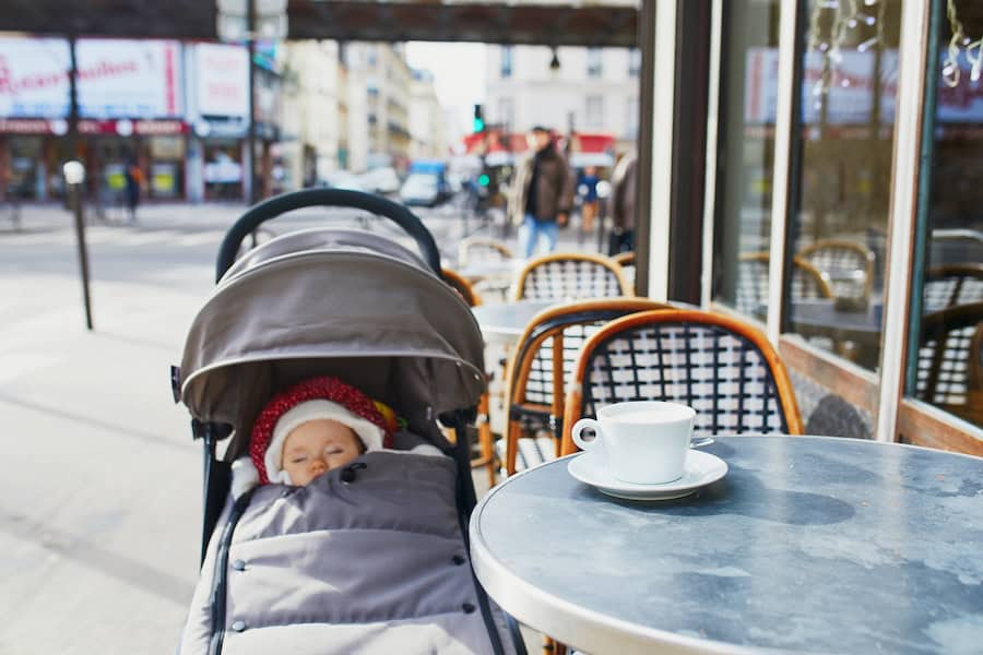 Baby girl sleeping in pram on outdoor terrace of Parisian street cafe with cup of hot coffee on the table.