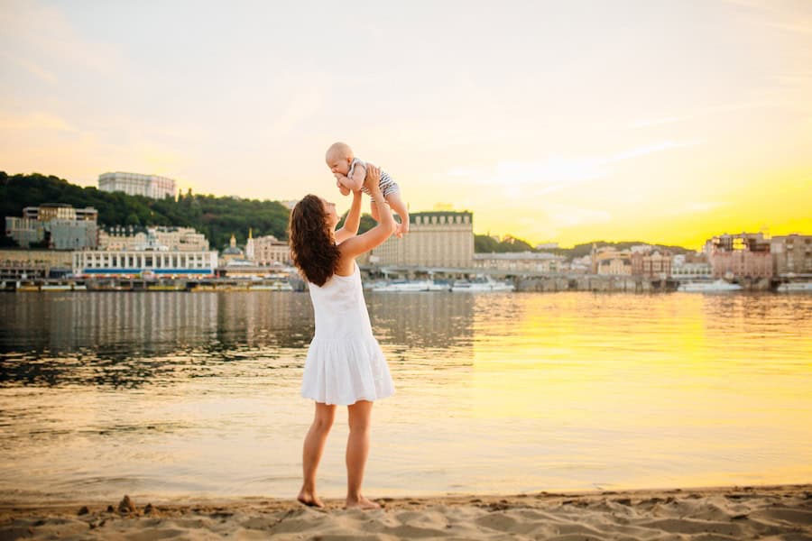 mother holding infant son high against a sunset and water background.