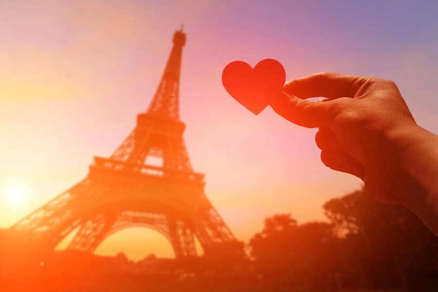 A silhouette of the Eiffel Tower in Paris against the sunset, while someone's hand holds a paper heart in the foreground